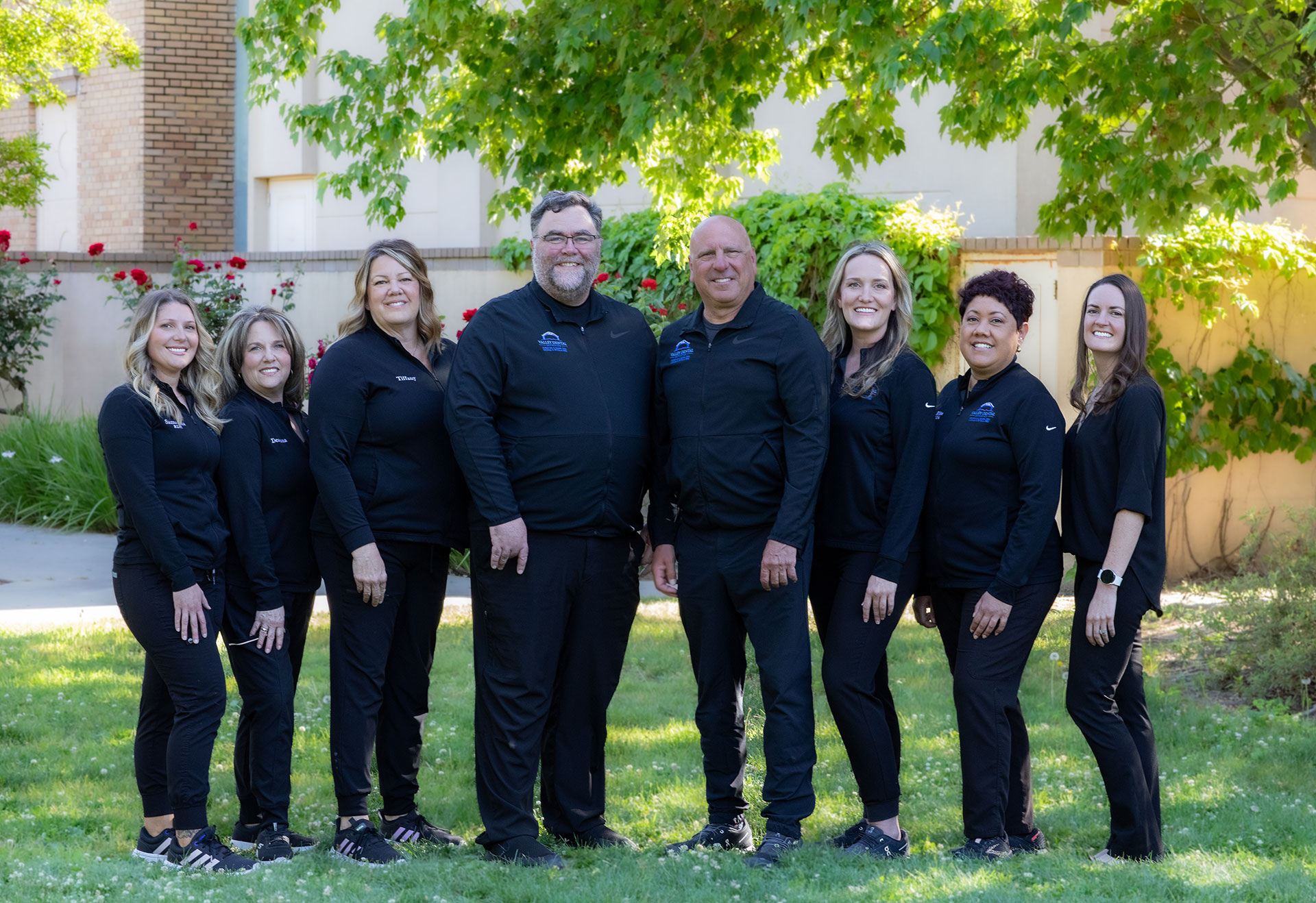 The image features a group of seven individuals, likely a team or organization, posing for a photograph outdoors. They are dressed in black shirts with visible logos and white pants, suggesting they may be part of an official uniformed group or sports team.