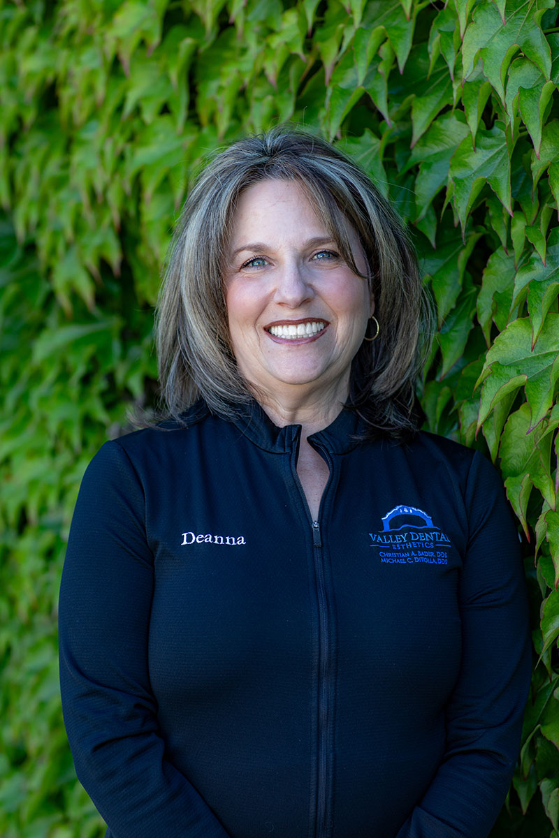 The image is a portrait of a woman standing outdoors, smiling gently at the camera. She has short hair, wears a dark jacket with a logo on it and a name tag, and has a professional appearance.