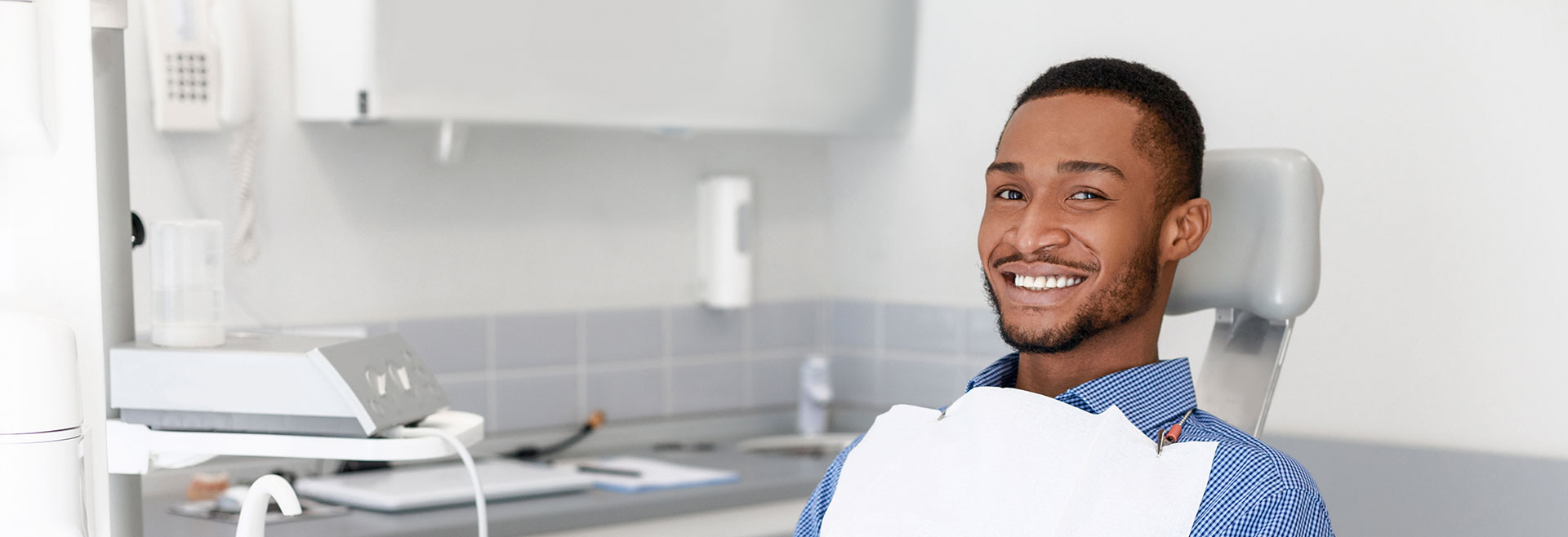 The image shows a person sitting in a dental chair with a broad smile, wearing a white apron and holding a toothbrush.