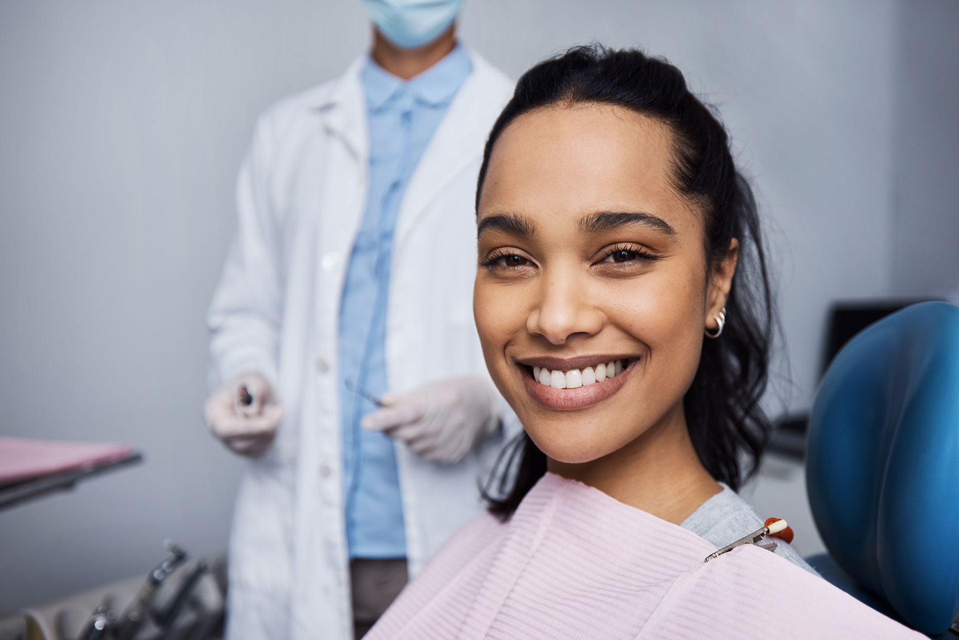 The image features a young woman sitting in a dental chair, smiling at the camera, with a dental hygienist standing behind her wearing protective gloves and a face mask.