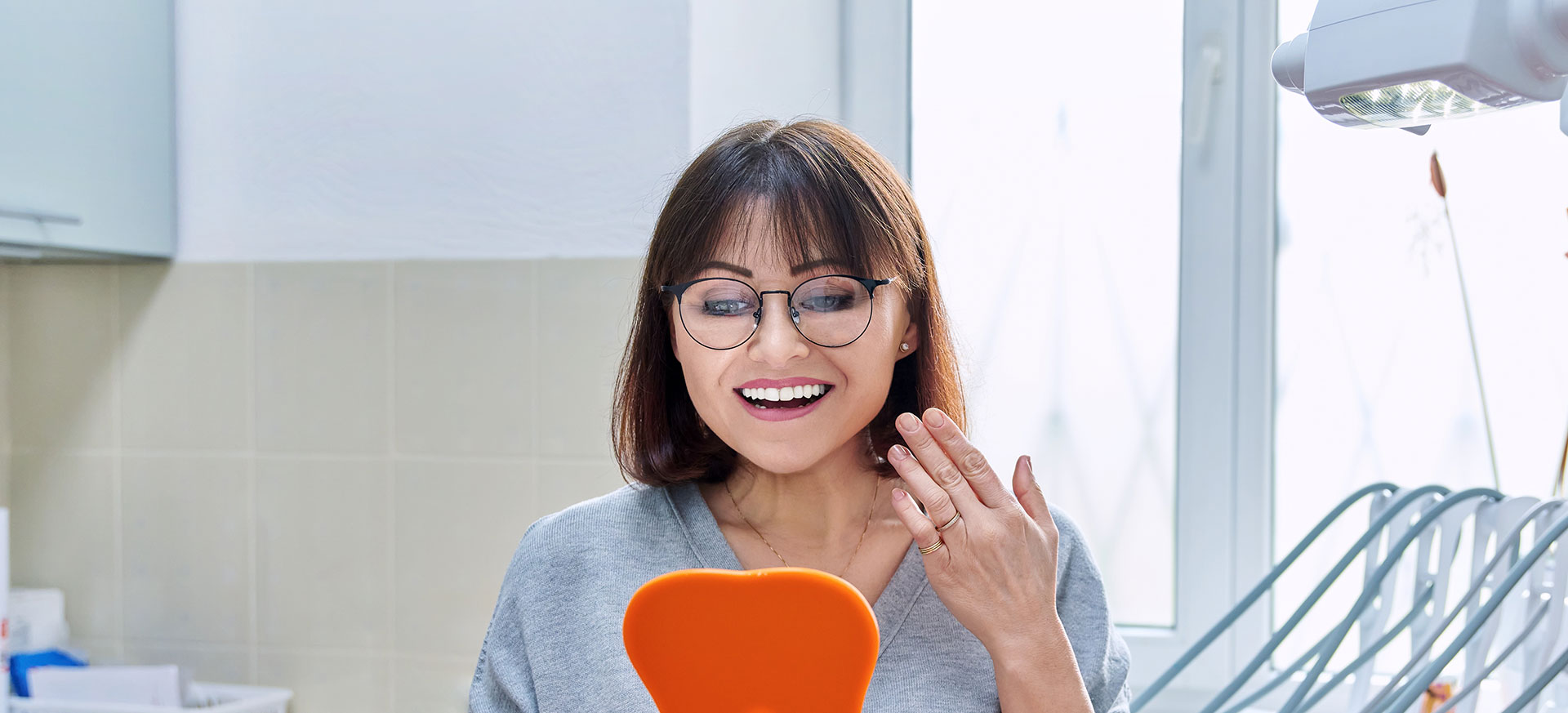 The image shows a woman with glasses, smiling and looking at her phone screen while standing in a kitchen environment.