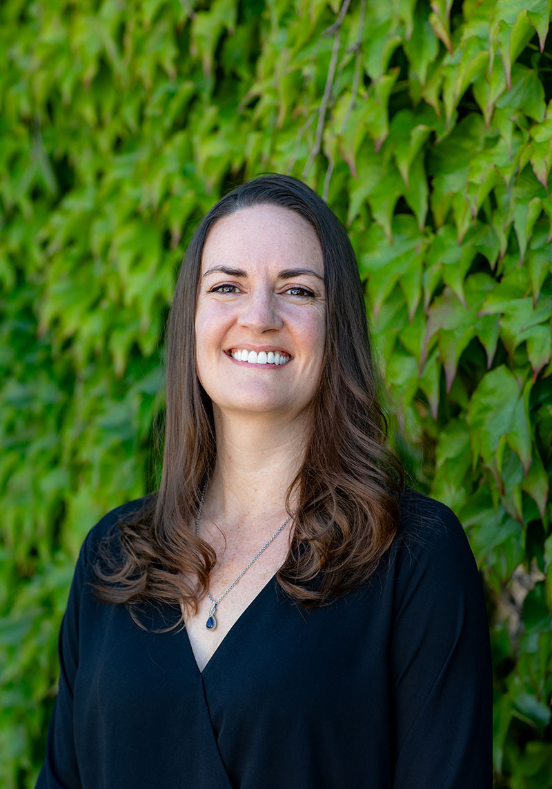 The image shows a smiling woman standing against a green wall with ivy, wearing a dark top and a necklace.