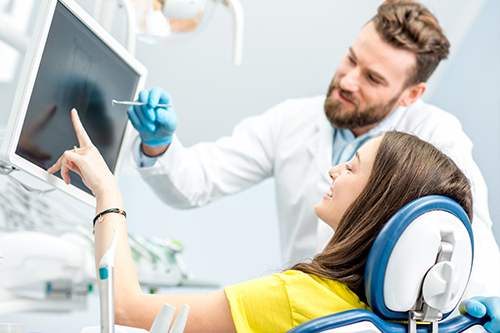 The image shows a medical professional, likely a dentist or oral surgeon, assisting a patient with a computer screen displaying what appears to be an X-ray image.