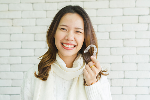 The image shows a smiling woman holding up an object that appears to be a dental appliance, with a brick wall in the background.